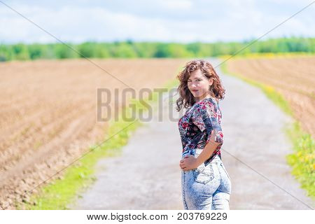 Young Woman Standing Posing In Jeans, Turned Back On Countryside Dirt Road By Brown Plowed Field Wit