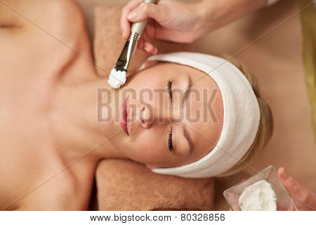 people, beauty, spa, cosmetology and skincare concept - close up of beautiful young woman lying with closed eyes and cosmetologist applying facial mask by brush in spa