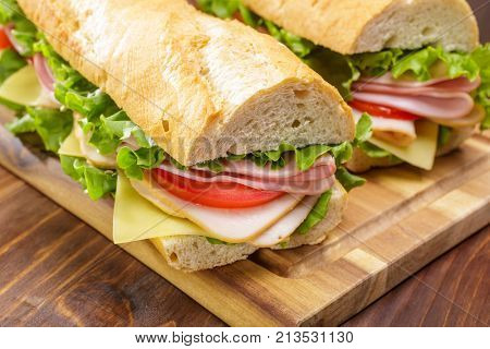 Baguette sandwich cut in half with ham,  turkey breast, cheese, lettuce and tomatoes on a cutting board closeup