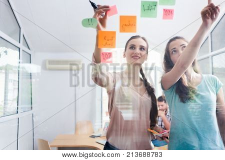 Two proficient female co-workers posting sticky notes in the interior of a shared office space for task prioritization and better time management