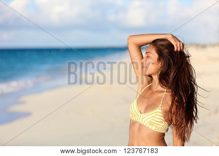 Carefree bikini woman enjoying sunset on beach. Beautiful relaxing Asian model caressing hair with closed eyes over the ocean on summer travel vacation in yellow swimsuit top in tropical destination.