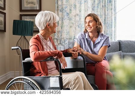 Mature woman comforting senior mom sitting on wheelchair at nursing home. Cheerful woman talking to old disabled mother at elder care centre. Loving caregiver taking care of elderly woman at home.