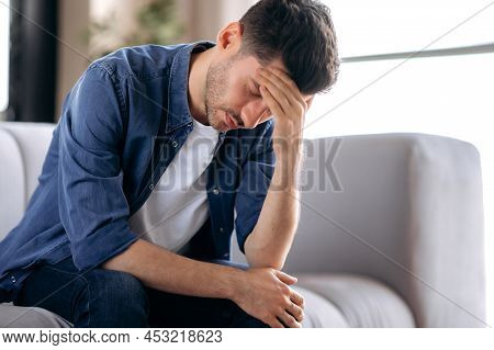 Upset Sick Stressed Young Caucasian Man In Casual Clothes Sits On Sofa In The Living Room, Bending O