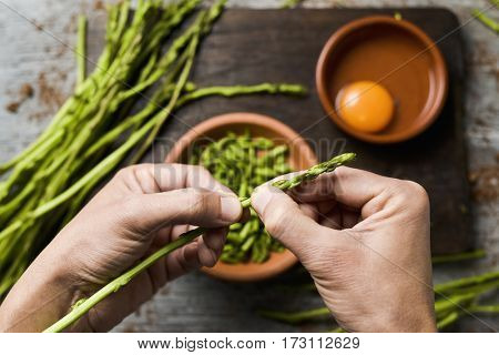 young man chopping wild asparagus with his hands and some earthenware bowls with chopped asparagus and a cracked egg, the ingredients to prepare an asparagus omelet, on a rustic wooden surface