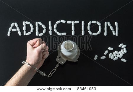 Drug addict or medical abuse concept with man handcuffed to a medicine bottle. Obsession to pharmaceutical substances or narcotics or anxiety pills. Addiction written with tablets on dark background.