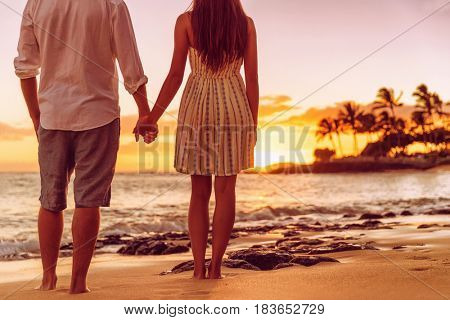 Beach couple watching sunset holding hands. People from behind relaxing enjoying summer travel vacation on tropical destination.