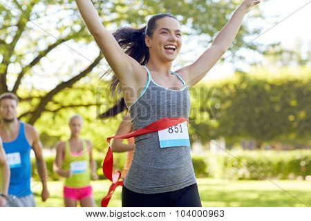 fitness, sport, victory, success and healthy lifestyle concept - happy woman winning race and coming first to finish red ribbon over group of sportsmen running marathon with badge numbers outdoors