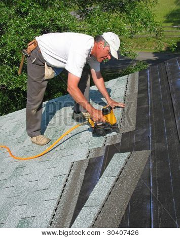 Carpenter uses nail gun to attach asphalt shingles to roof