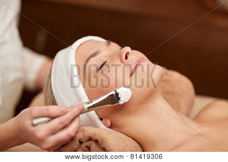 people, beauty, spa, cosmetology and skincare concept - close up of beautiful young woman lying with closed eyes and cosmetologist applying facial mask by brush in spa