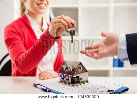 Closeup of smiling woman realtor giving key of new apartment to client. Female house agent sitting at the desk with house miniature and plan.