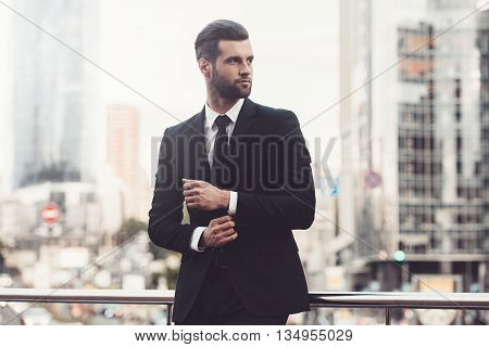 Modern businessman. Confident young man in full suit adjusting his sleeve and looking away while standing outdoors with cityscape in the background