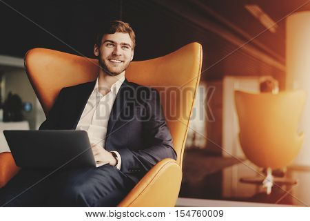 Young smiling successful man entrepreneur in formal business suite with a beard sitting on orange armchair with laptop in luxury office interior