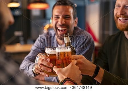 Happy mid adult friends clinking with beer mugs in pub. Three cheerful guys drinking draft beer, celebrating meeting and smiling. Laughing young men enjoying cold pint of beer during night at bar. 