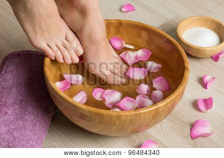 Closeup shot of a woman feet dipped in water with petals in a wooden bowl. Beautiful female feet at spa salon on pedicure procedure. Shallow depth of field with focus on feet.
