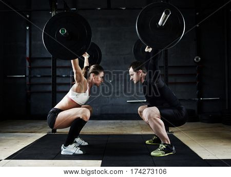 Motivational side view image of young man and woman performing overhead squats with huge heavy barbells looking at each other