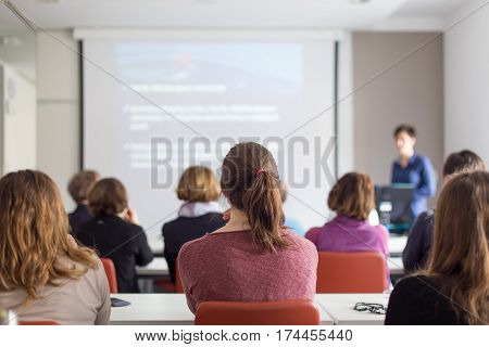 Female speaker giving presentation in lecture hall at university workshop. Rear view of unrecognized participants listening to lecture and making notes. Scientific conference event.