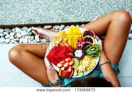 Girl relaxing and eating fruit plate by the hotel pool. Exotic summer diet. Photo of legs with healthy food by the poolside, top view from above. Tropical beach lifestyle.