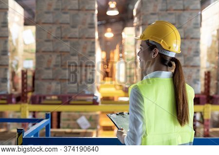 Side view of asian female warehouse worker with archboard flip chart look inventory with dustrubution warehouse in background. Reopening business warehouse technology and logistic concept.