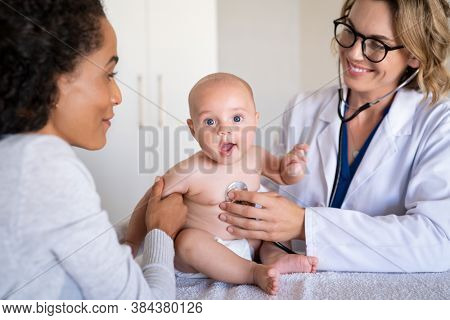 Female smiling doctor examining little baby with stethoscope in clinic. Portrait of a cute little boy checked by doctor with his mother. Pediatrician examining baby boy with a stethoscope. 