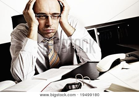 Worried businessman sitting at office desk full with books and papers being overloaded with work.