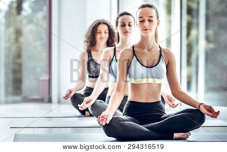 Group Of Young People Doing Yoga Sitting On Mats. Three People Practicing Meditation And Yoga On Cla