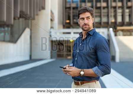 Man using smartphone outdoor. Portrait of young businessman using mobile phone while looking away on the street. Thoughtful business man in blue formal clothing using cellphone outside office building