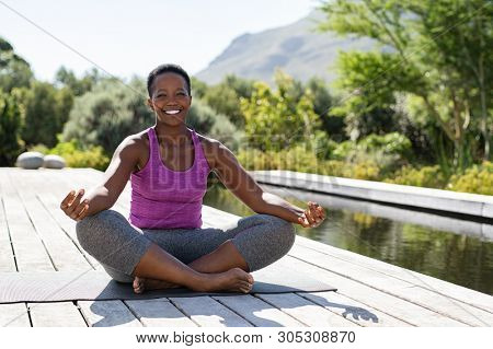 Portrait of smiling woman in lotus position near swimming pool looking at camera. Healthy black mature woman meditating and sitting in lotus pose on yoga mat. Happy lady in zen pose doing meditation.