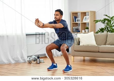 sport and healthy lifestyle concept - indian man with fitness tracker exercising and doing squats at home