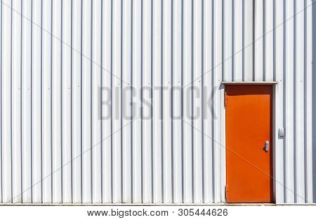 Industrial Building Facade With A Metal Orange Door And A White Aluminum Sheet Wall. White Warehouse