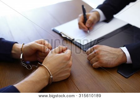 Police Officer Interrogating Criminal In Handcuffs At Desk Indoors