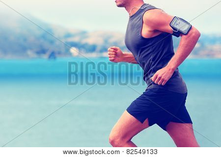 Cardio runner running listening smartphone music. Unrecognizable body jogging on ocean beach or waterfront working out with heart rate monitor app device and earphones in summer.