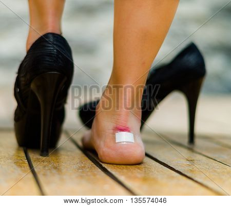 High heels hurts very often, feet with white little patch on ankle, one feet on the floor and other with black shoe.