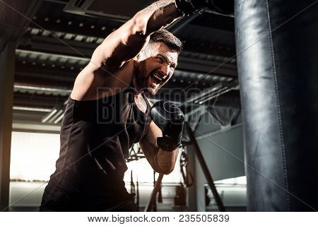 Bearded Male Boxer Training With Punching Bag In Dark Sports Hall. Young Tattoed Boxer Training On P