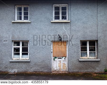 An Old Abandoned Grey Derelict House On A Residential Street With Dirty Broken Windows And A Boarded