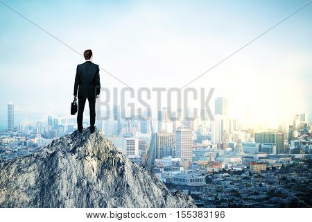 Back view of young man in suit and with briefcase in hand standing on mountain top. City background with copy space. Leadership concept