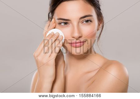 Healthy fresh girl removing makeup from her face with cotton pad. Beauty woman cleaning her face with cotton swab pad isolated on grey background. Skin care and beauty concept.