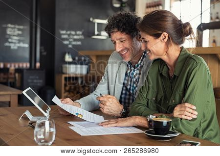 Portrait of mature business man and casual businesswoman sitting in cafe and discussing sales graph. Group of two middle aged coworkers working comparing forecasting graphics. Happy businesspeople.