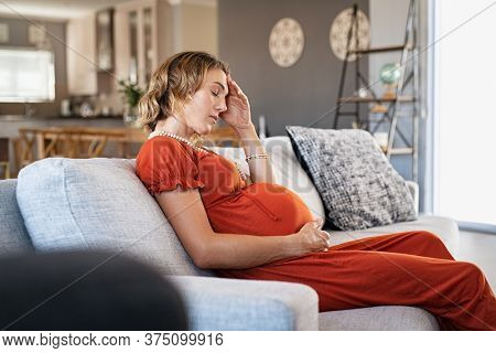 Exhausted middle aged pregnant woman suffering headache sitting on couch in the living room. Stressed young pregnant woman with terrible migraine. Expecting mother holding belly bump feeling worrying.