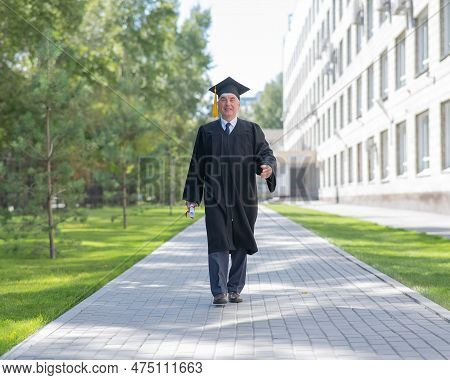 An Old Man In A Graduation Gown Walks Outdoors And Holds A Diploma.