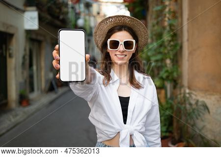 Glad Young Millennial European Female Tourist In Sunglasses And Hat Show Phone With Blank Screen In 