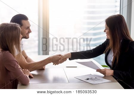 Young smiling couple visiting real estate agency shaking hands with realtor, purchasing renting flat or house, apartment buyers making deal, family couple and broker handshaking after signing papers