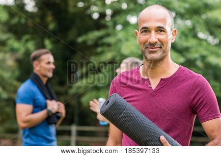 Smiling mature man holding yoga mat and looking at camera. Portrait of a happy mixed race man with yoga mat at park after fitness exercise. Healthy positive senior man with people in background.