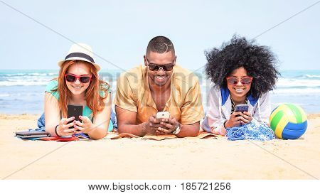 Row of multicultural friends using phone lying on beach - Happy african and american students on summer vacation texting messages are looking addicted at mobile technology and social media post -