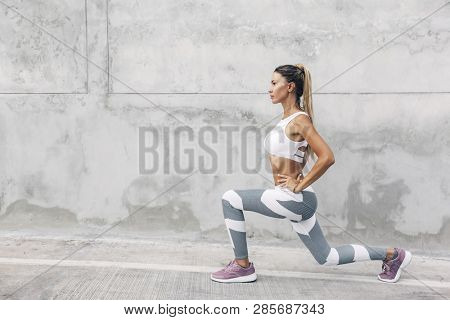 Fitness woman in sportswear doing squat exercise on the city street over gray concrete background. Outdoor sports clothing and shoes, urban style.