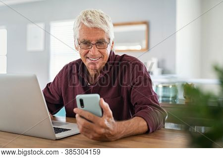 Mature man using smartphone in living room. Happy senior businessman working at home while messaging on mobile phone. Happy smiling old man using social media network technology and feeling excited.