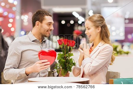 love, romance, valentines day, couple and people concept - happy young man with red flowers giving present to smiling woman at cafe in mall