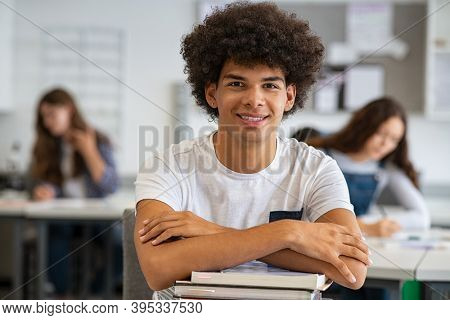 Portrait of happy african university student  in class looking at camera. Smiling young man at college leaning on stack of books with classmates in background. Satisfied african guy at college.