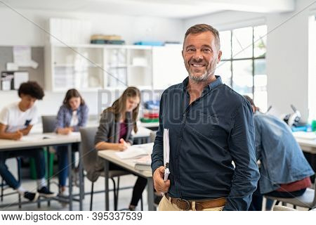 Portrait of mature teacher looking at camera with copy space. Happy mid adult lecturer at classroom standing after giving lecture. Satisfied high school teacher smiling while his students studying.