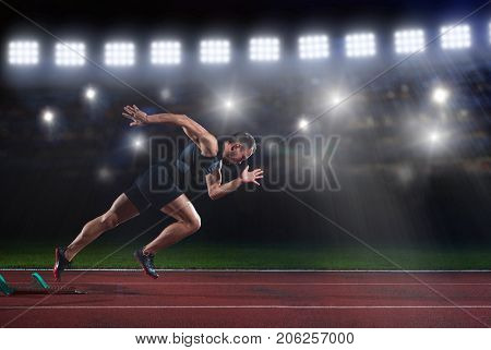 Sprinter leaving starting blocks on the running track in front of big modern stadium with lights and flares. Explosive start.