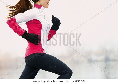 Winter running athlete woman on cold run jogging fast with speed and sprint on outside workout wearing warm clothing gloves, winter tights and wind jacket in snow weather.